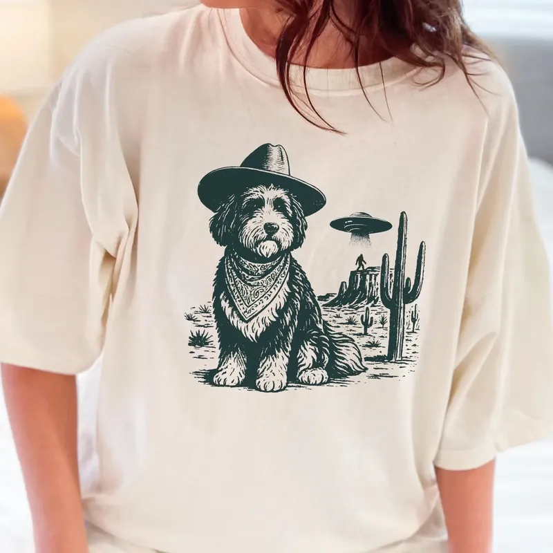 Cream tee printed with a sketched doodle in a cowboy hat and bandana, cacti and a UFO in the desert behind